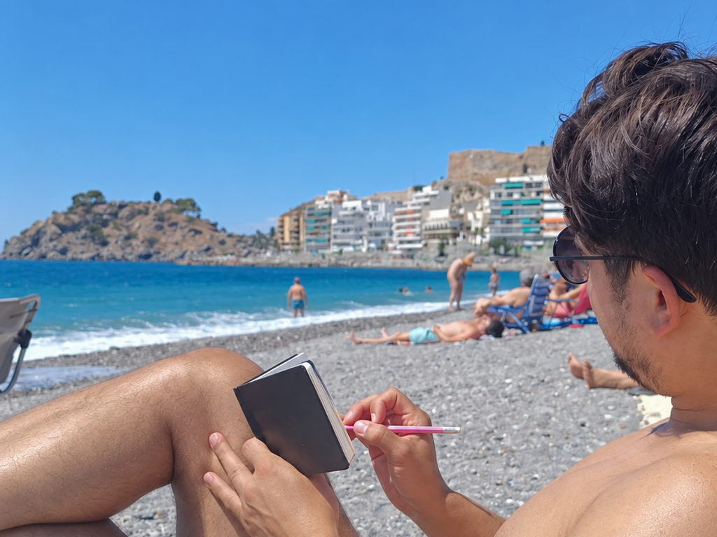 Cuaderno con una acuarela de una persona sentada frente al mar, sostenido ante la playa de Almuñécar con bañistas y rocas al fondo.