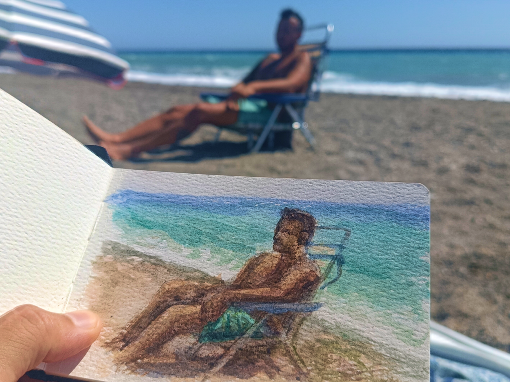 Cuaderno sobre una toalla con una acuarela de una figura bajo una sombrilla azul, sombra violeta en la arena y mar al fondo.