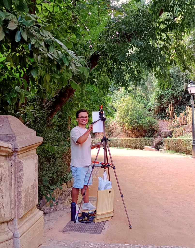 Yohan Pixone sonríe junto a su caballete, con una paleta en la mano, bajo los árboles del entorno de la Alhambra.