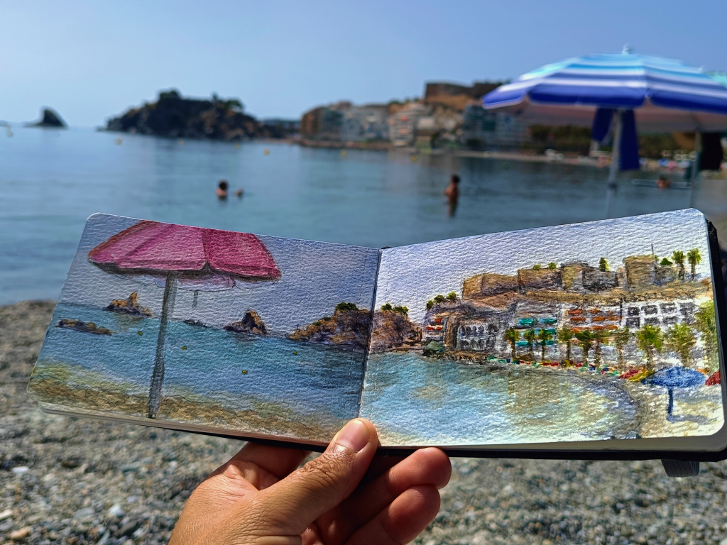 Cuaderno abierto con una acuarela de Yohan Pixone de sombrillas, mar y edificios de Almuñécar, sostenido frente a la costa.