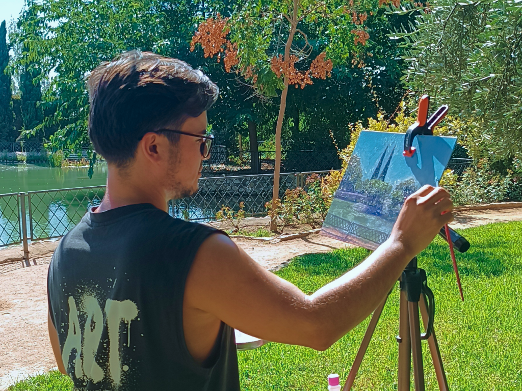 Yohan Pixone pintando descalzo sobre el césped junto al estanque del Parque Federico García Lorca, con el caballete bajo un árbol.